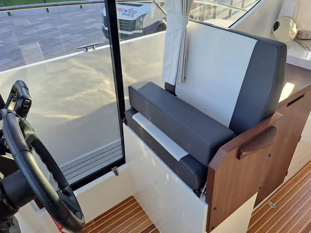 A modern boat interior featuring a steering wheel, seating area with grey and black upholstery, and striped wooden flooring.